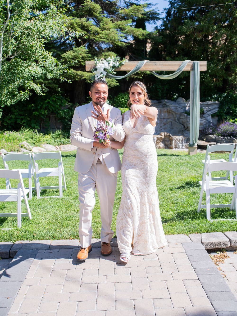 A couple celebrates during a backyard wedding ceremony with white chairs set up on a stone patio surrounded by greenery.