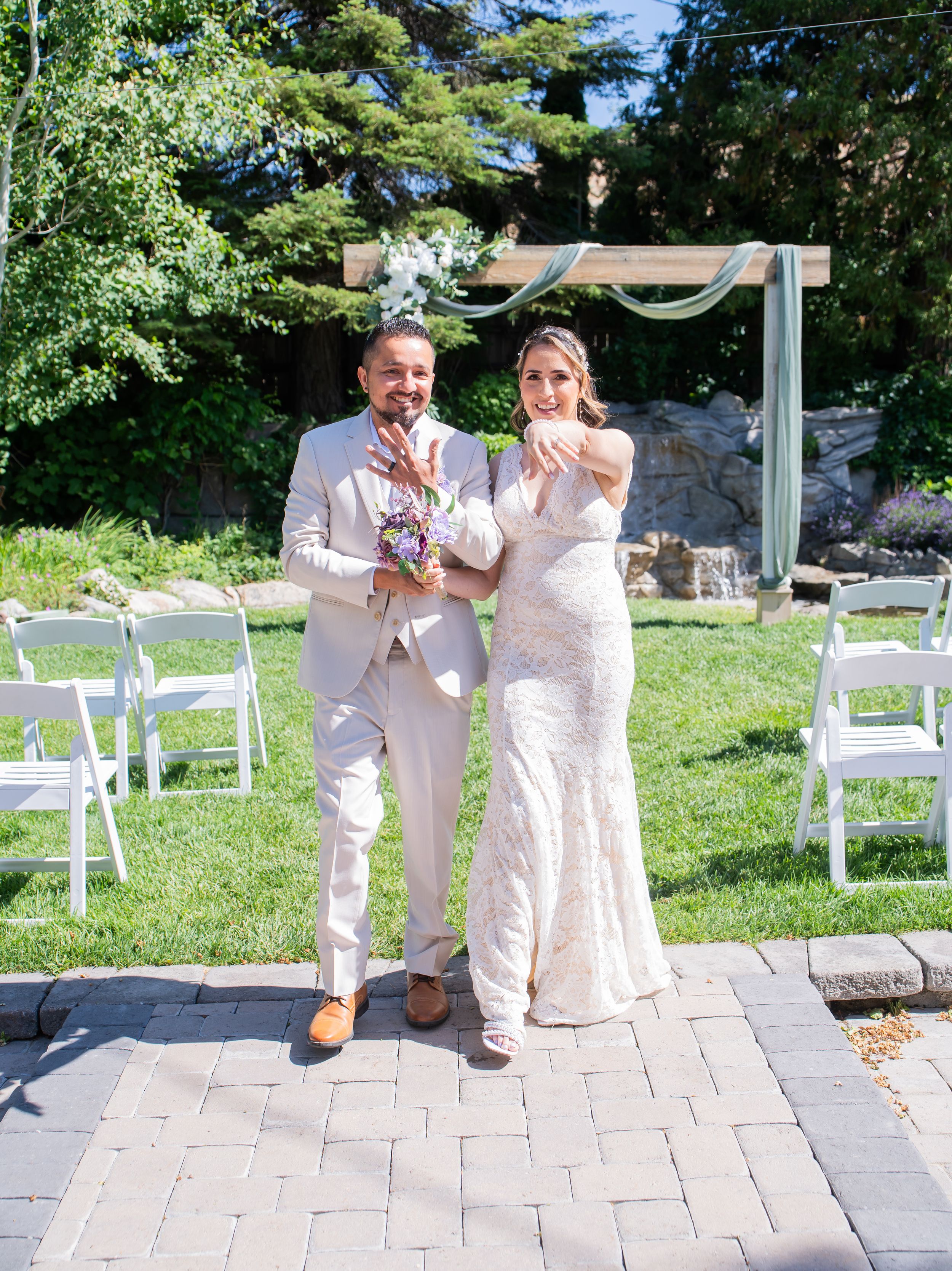 A couple celebrates during a backyard wedding ceremony with white chairs set up on a stone patio surrounded by greenery.