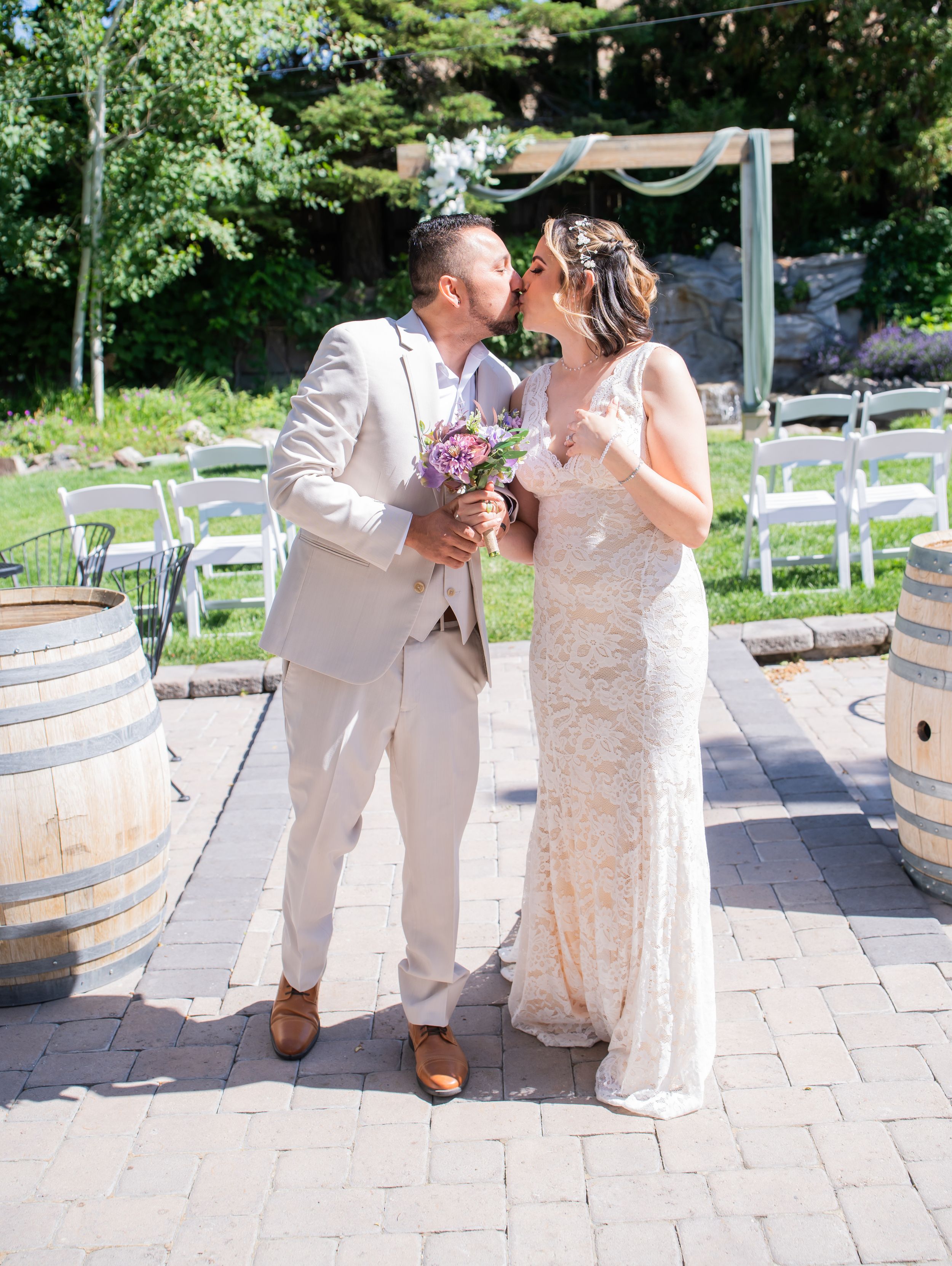 A romantic outdoor ceremony moment with two people in white outfits embracing and kissing beside rustic wine barrels.