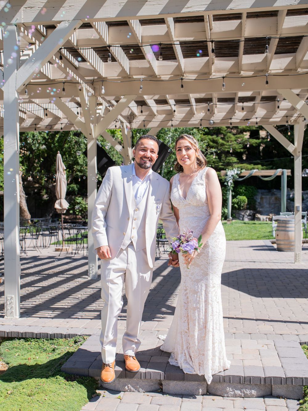 Wedding couple sharing joyful moments under an elegant white pergola in a sun-lit outdoor venue.