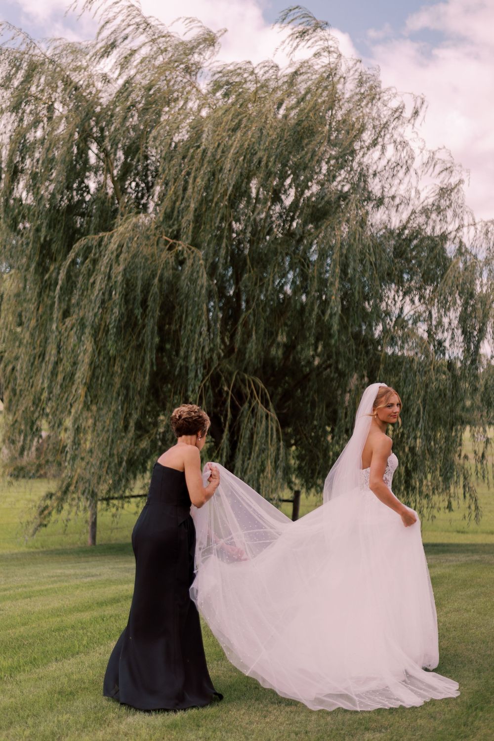 Bridesmaid in black dress helps arrange bride's flowing train under weeping willow tree.