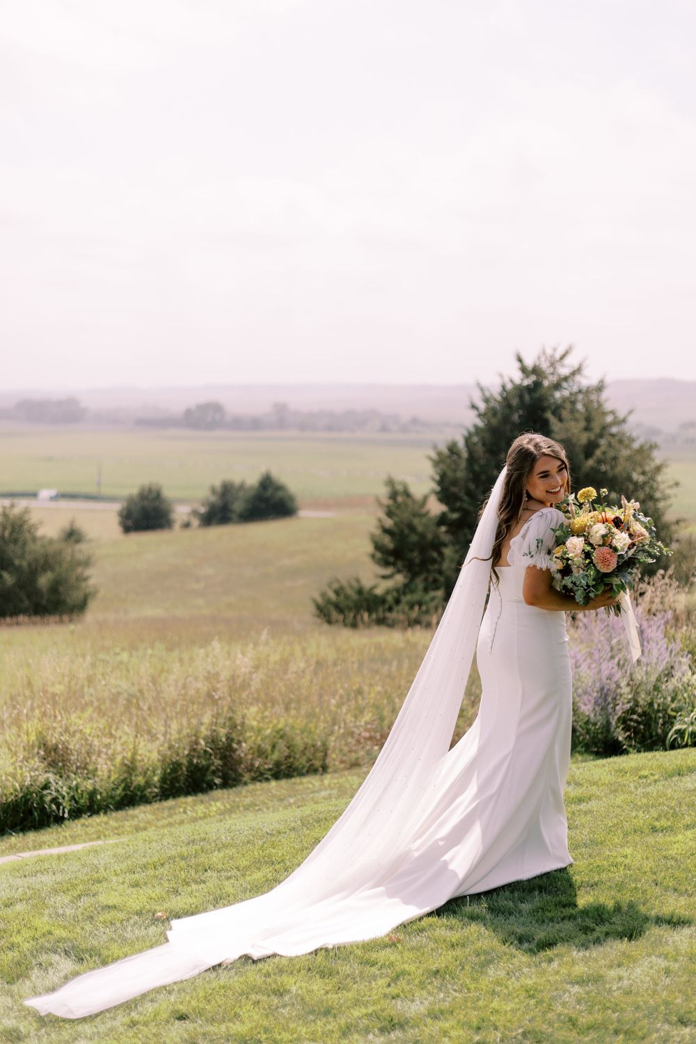 Bride in flowing white dress holding bouquet on hilltop overlooking countryside.