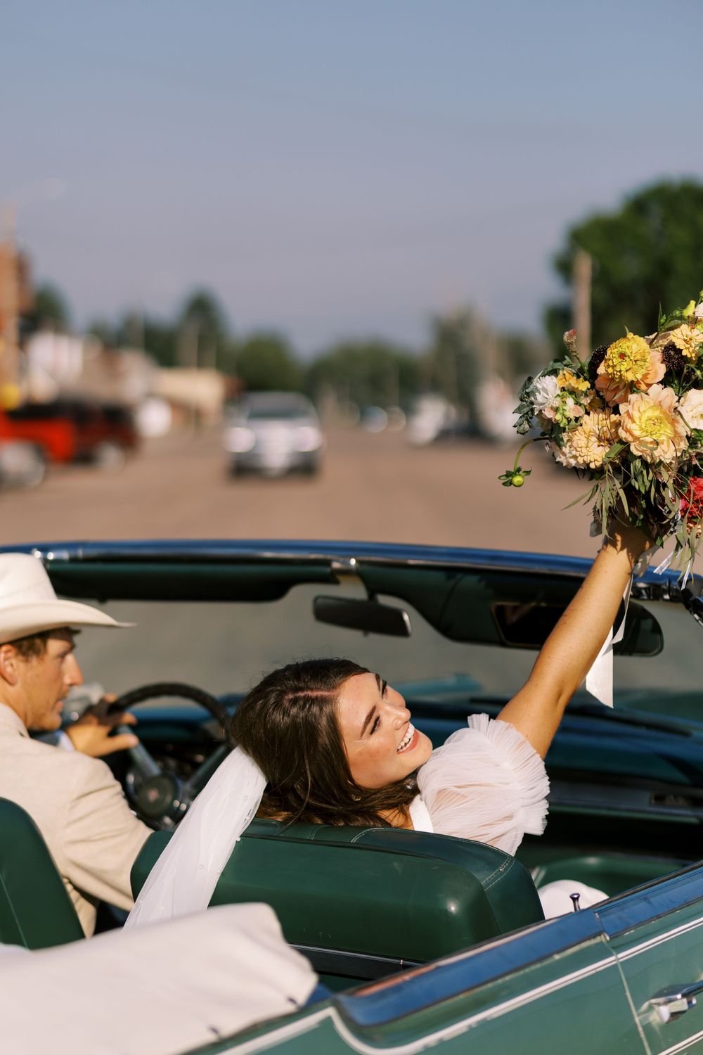 Newlyweds celebrating in vintage convertible with bouquet held high.