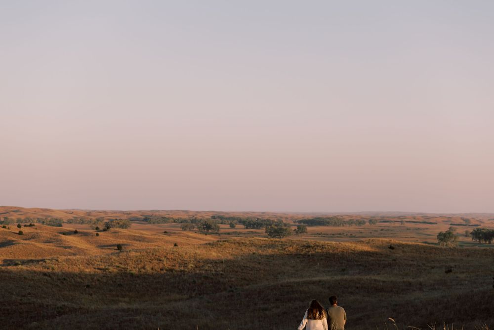 Two people sitting on hill overlooking expansive prairie landscape at sunset.