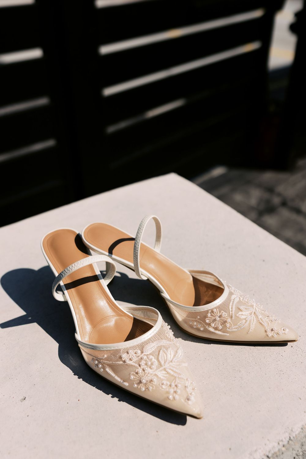 Elegant beige bridal heels with delicate floral embroidery casting shadows in natural sunlight on concrete surface.