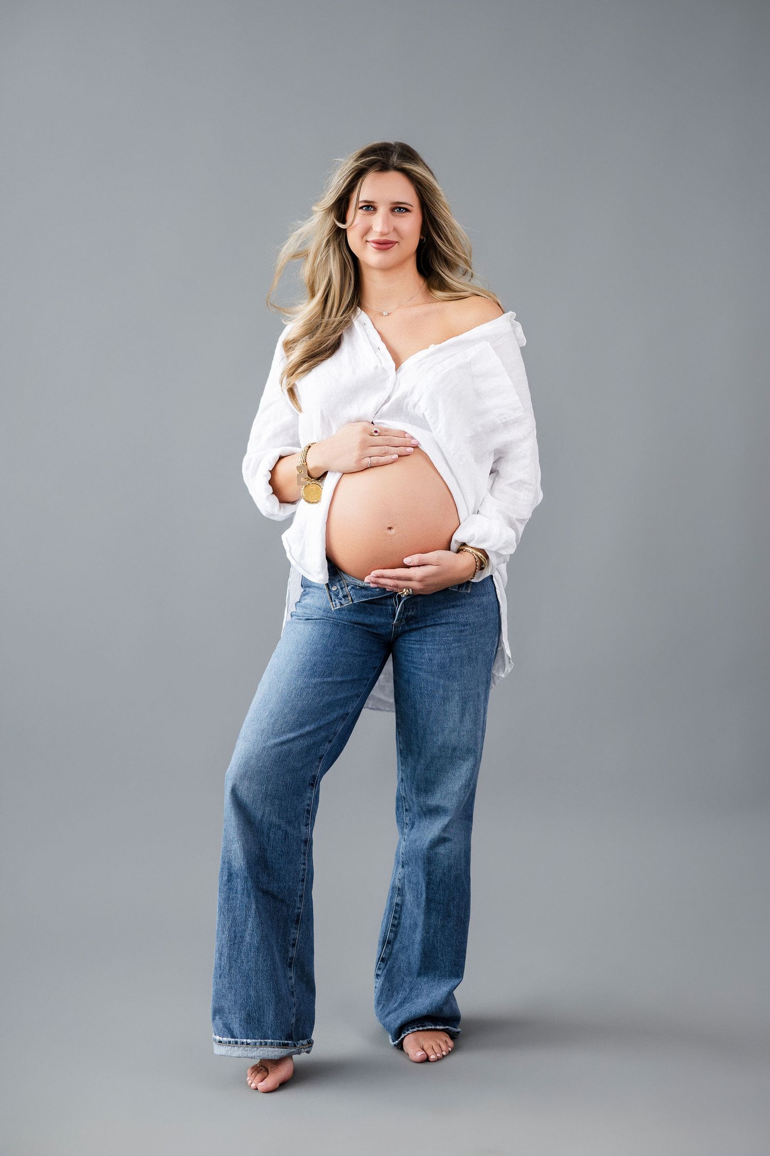 Maternity portrait showing pregnant woman in white button-down shirt and jeans against gray background.