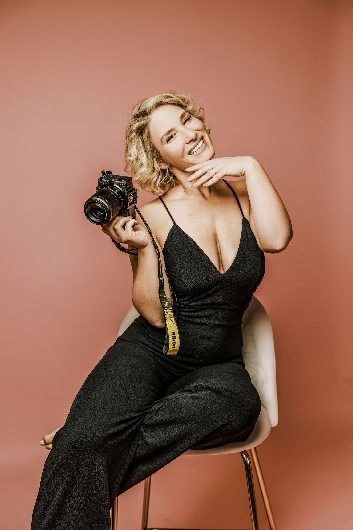 Professional photographer in elegant black jumpsuit poses with DSLR camera against coral backdrop in studio setting.