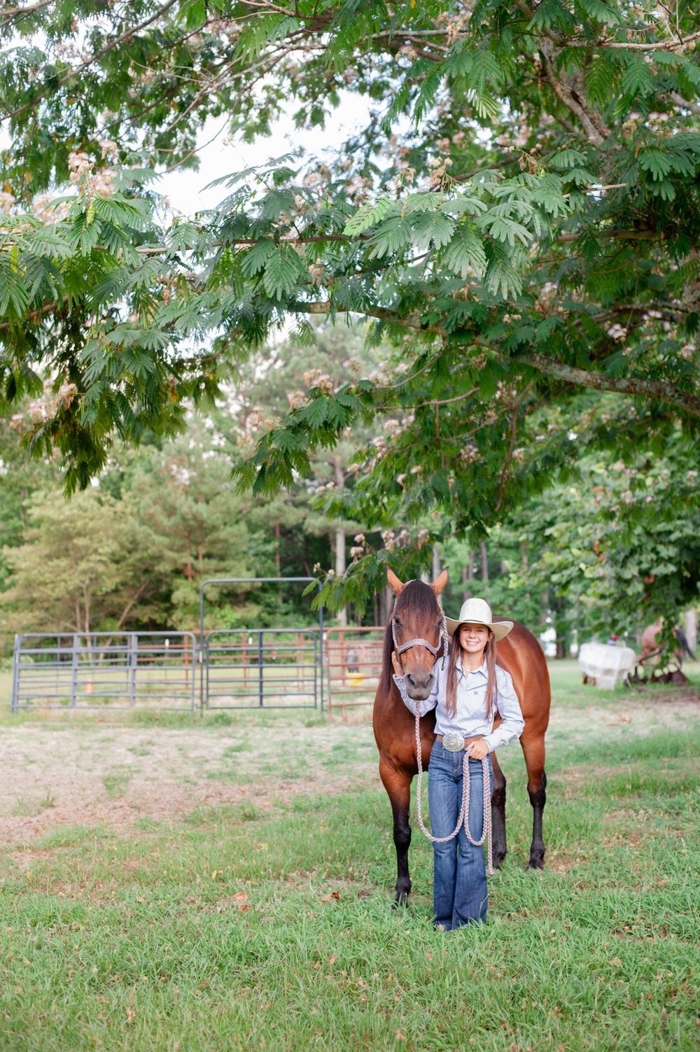 A person in western wear stands beside a tall brown horse in a pastoral farm setting.
