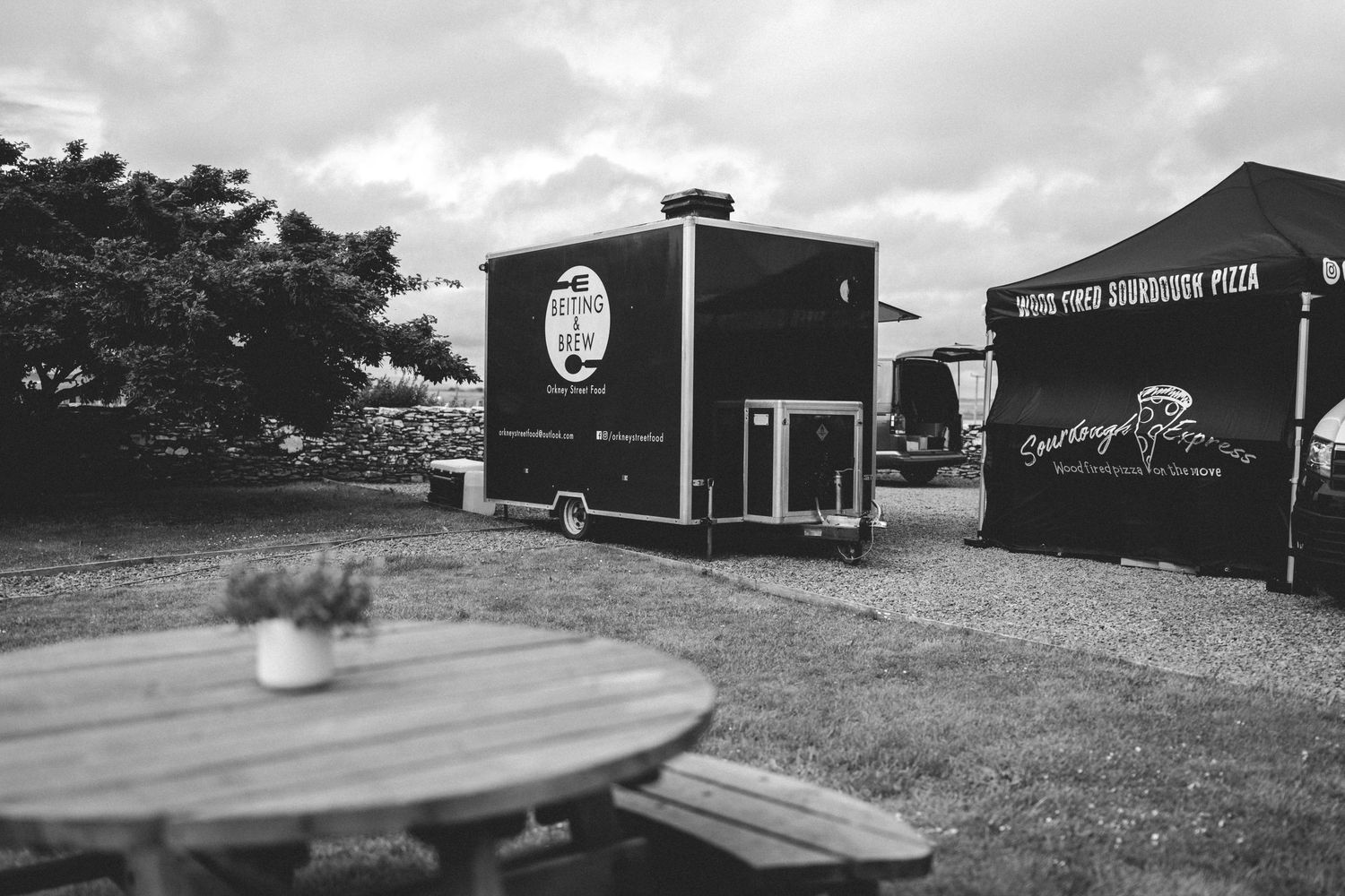 A black and white photo of food trucks and outdoor seating area with wooden tables on a cloudy day.