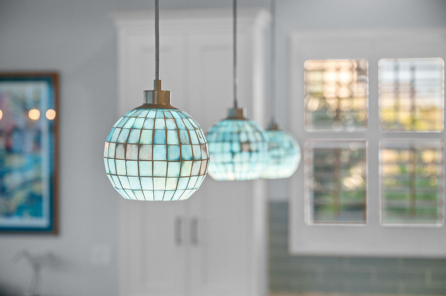 Three turquoise glass globe pendant lights hang in a bright room with windows and blurred background decor.