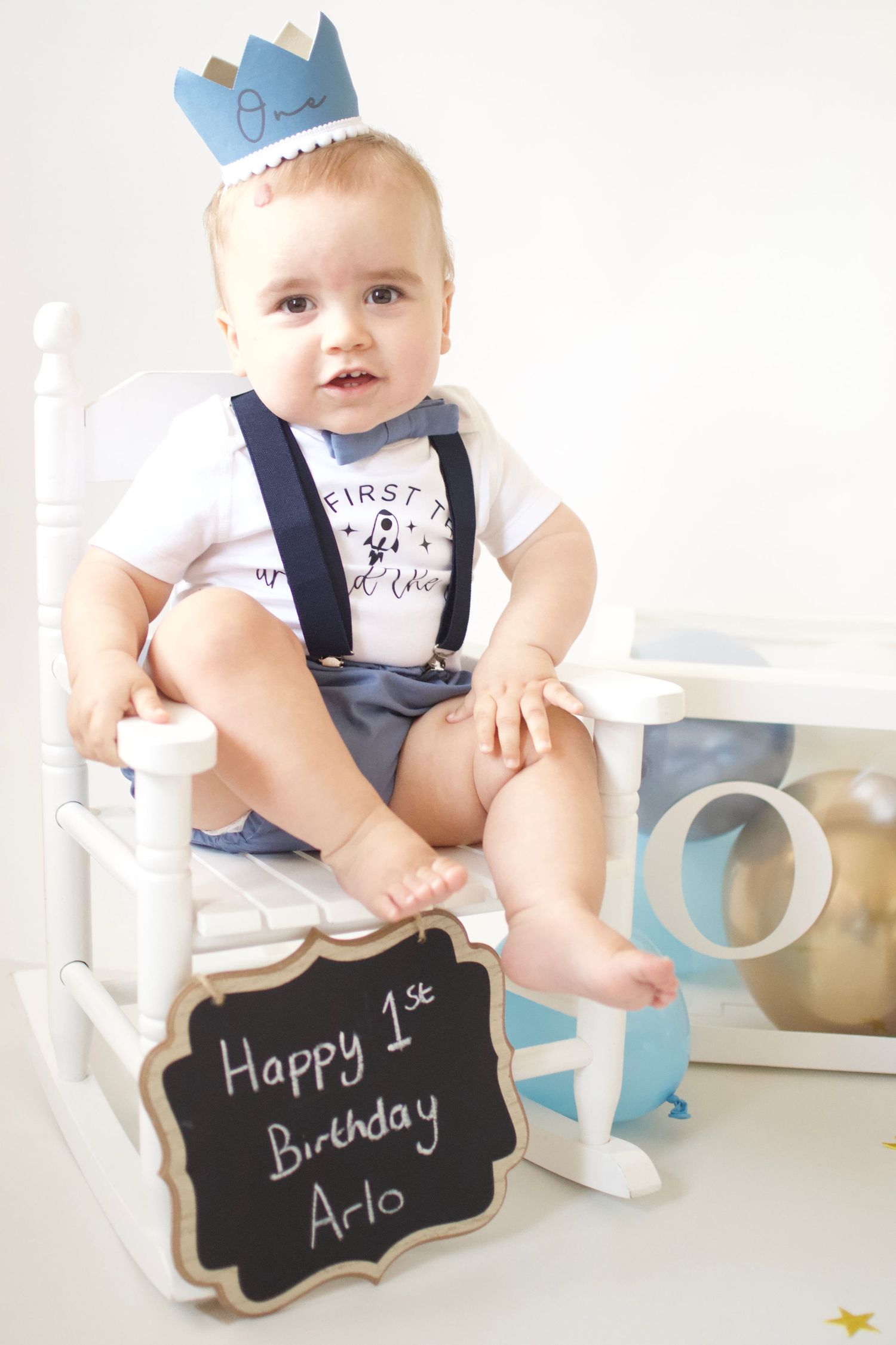 Baby in navy suspenders poses with first birthday chalkboard signs.