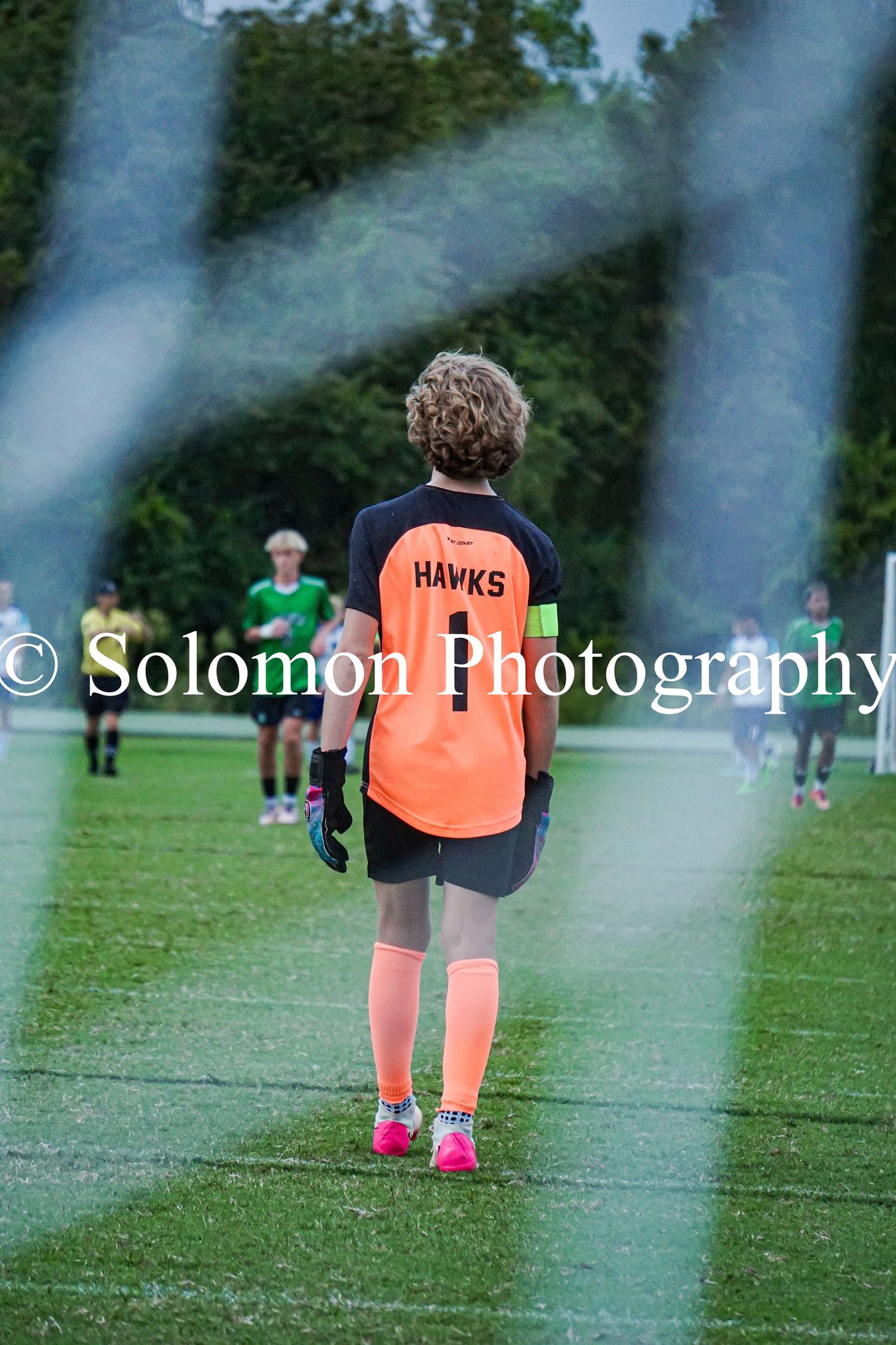 Hawks goalkeeper in orange jersey stands ready at goal during a soccer match by Solomon Photography.