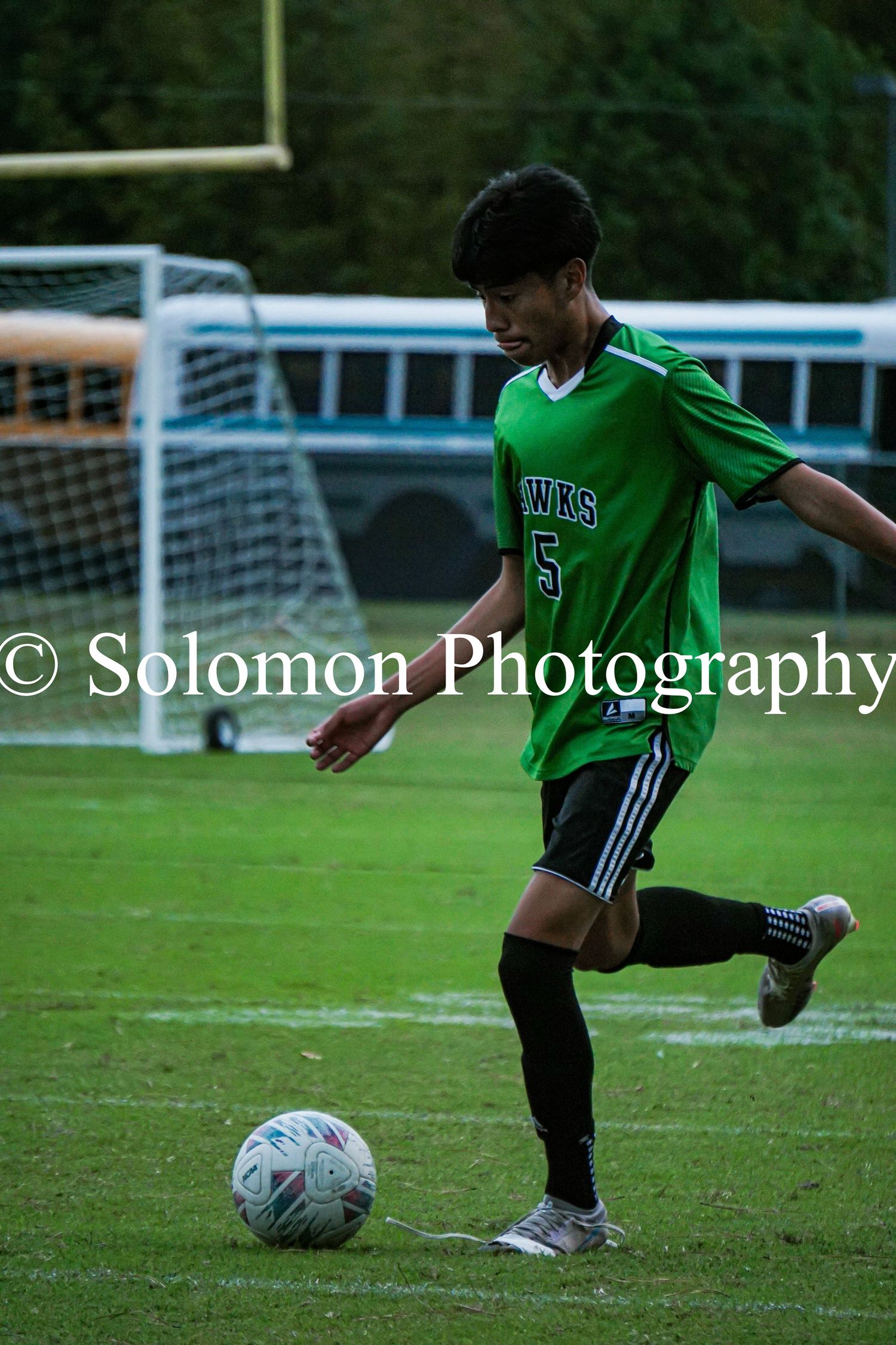 Soccer player in green Hawks #5 jersey dribbles ball on grass field.