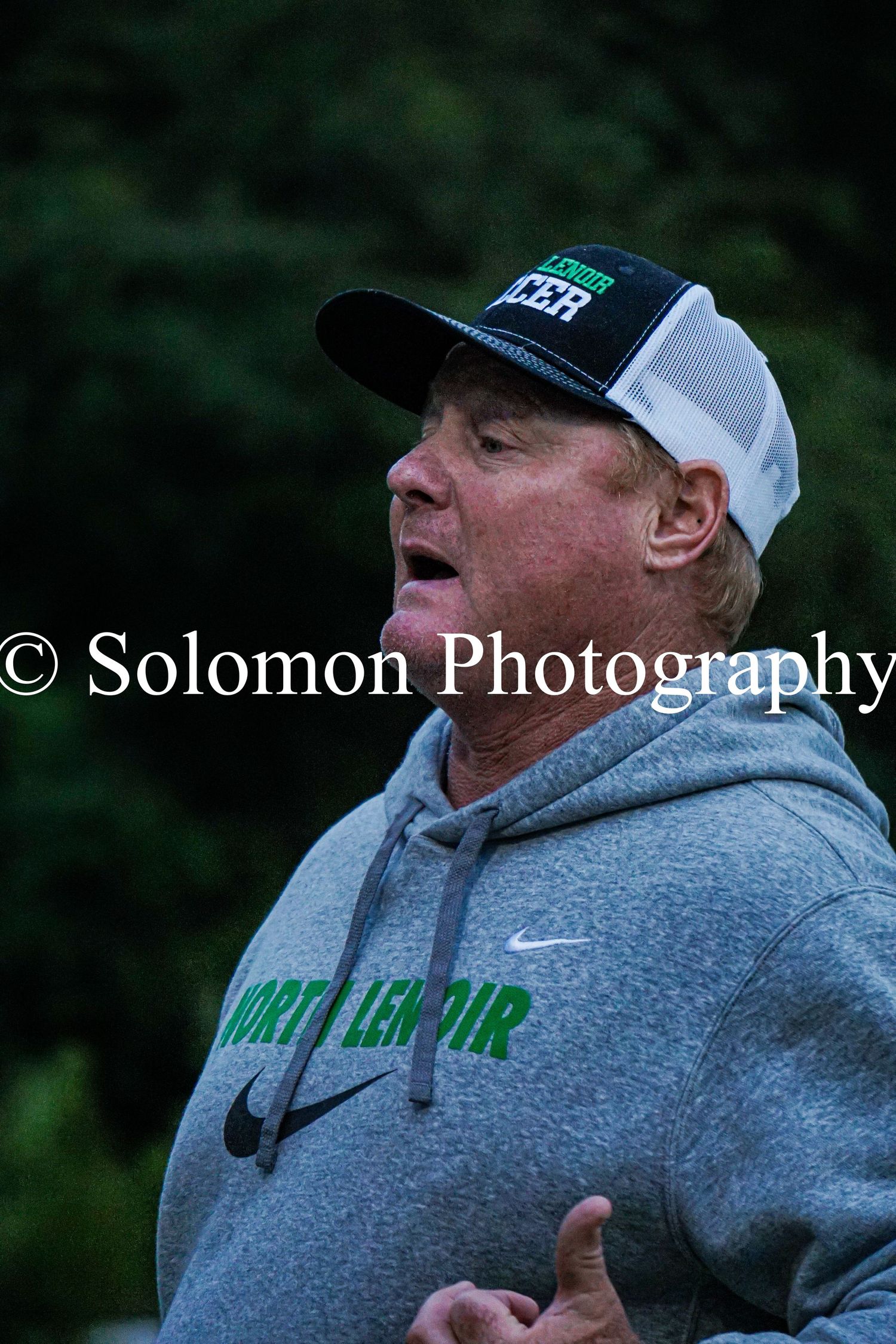 North Lenoir soccer coach in cap and gray hoodie shouts instructions during game.