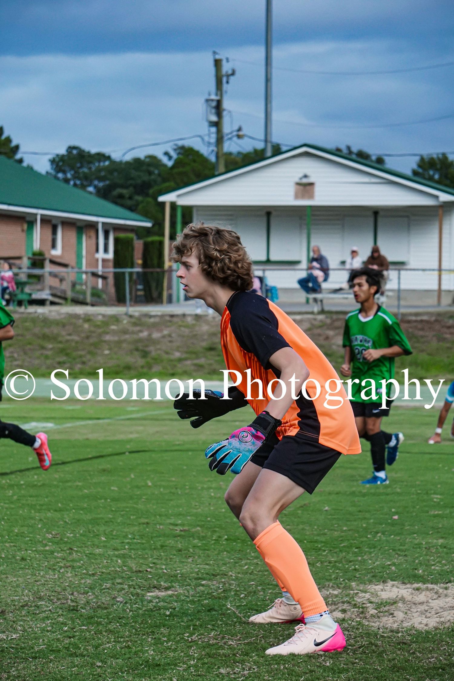 A goalkeeper in an orange uniform runs across a green soccer field during a match.