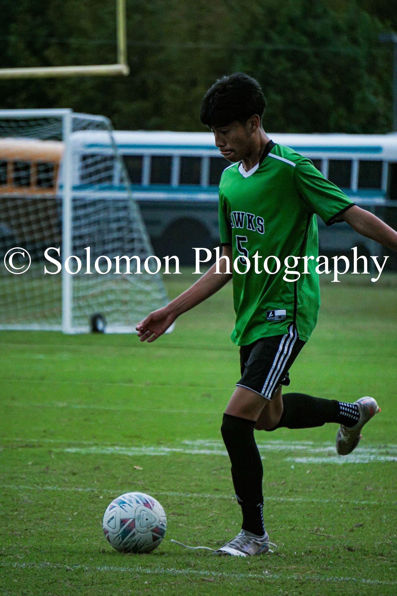 A Hawks soccer player in green dribbles a ball on the field during a game.