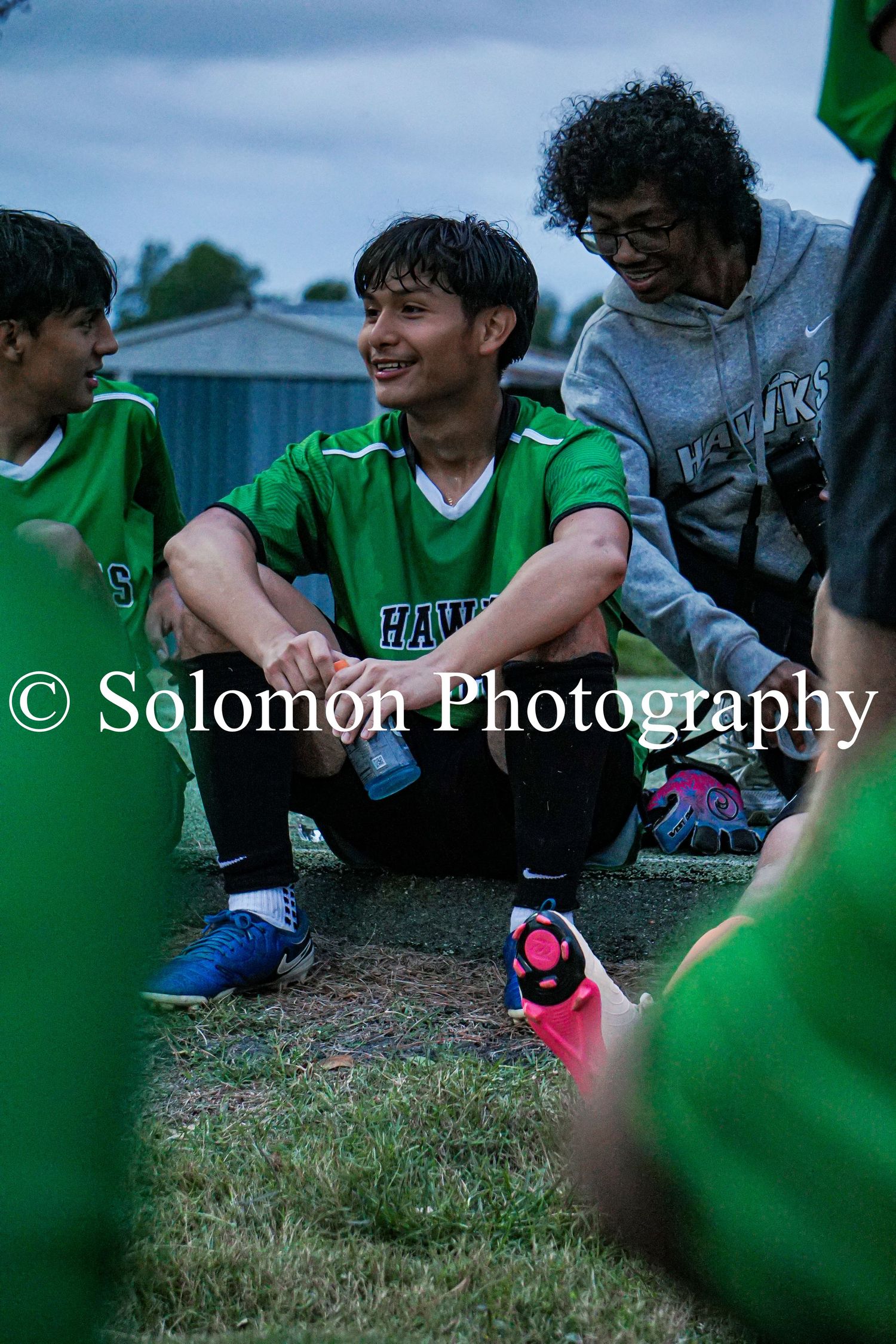 Soccer players in green Hawks jerseys sit on grass during a team huddle at an outdoor match.