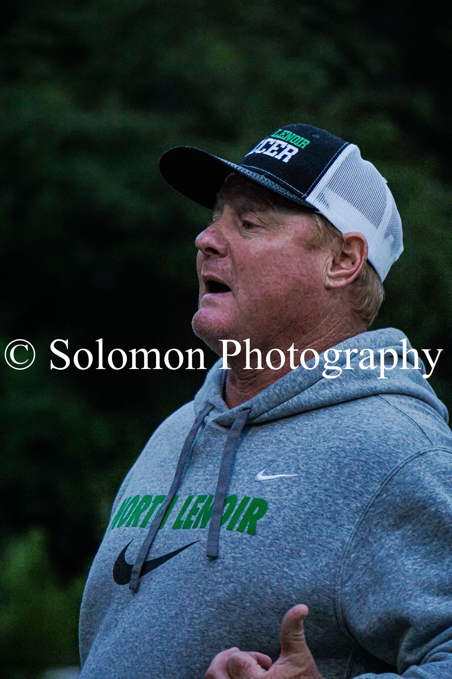 A North Lenoir soccer coach in a gray hoodie and green cap shouts instructions on the sideline.