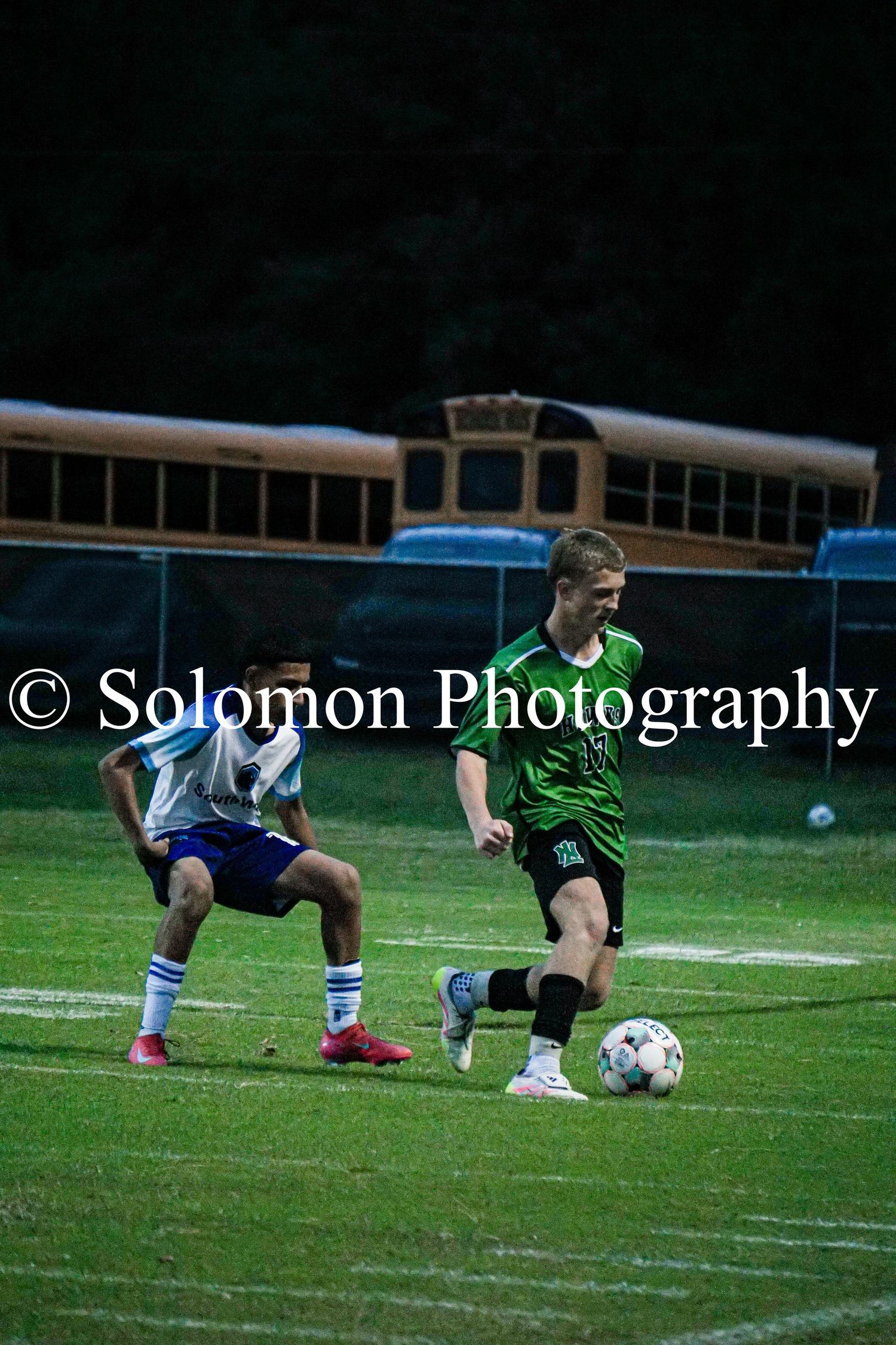 A North Lenoir soccer player in green dribbles past a defender near school buses.
