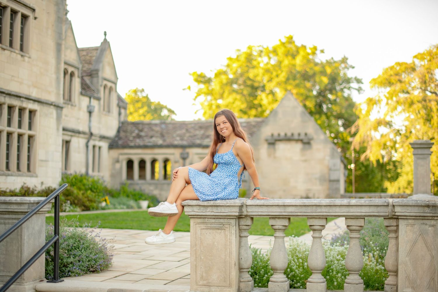 A person in a blue dress sits on a stone railing near a Gothic-style building on a sunny summer afternoon.