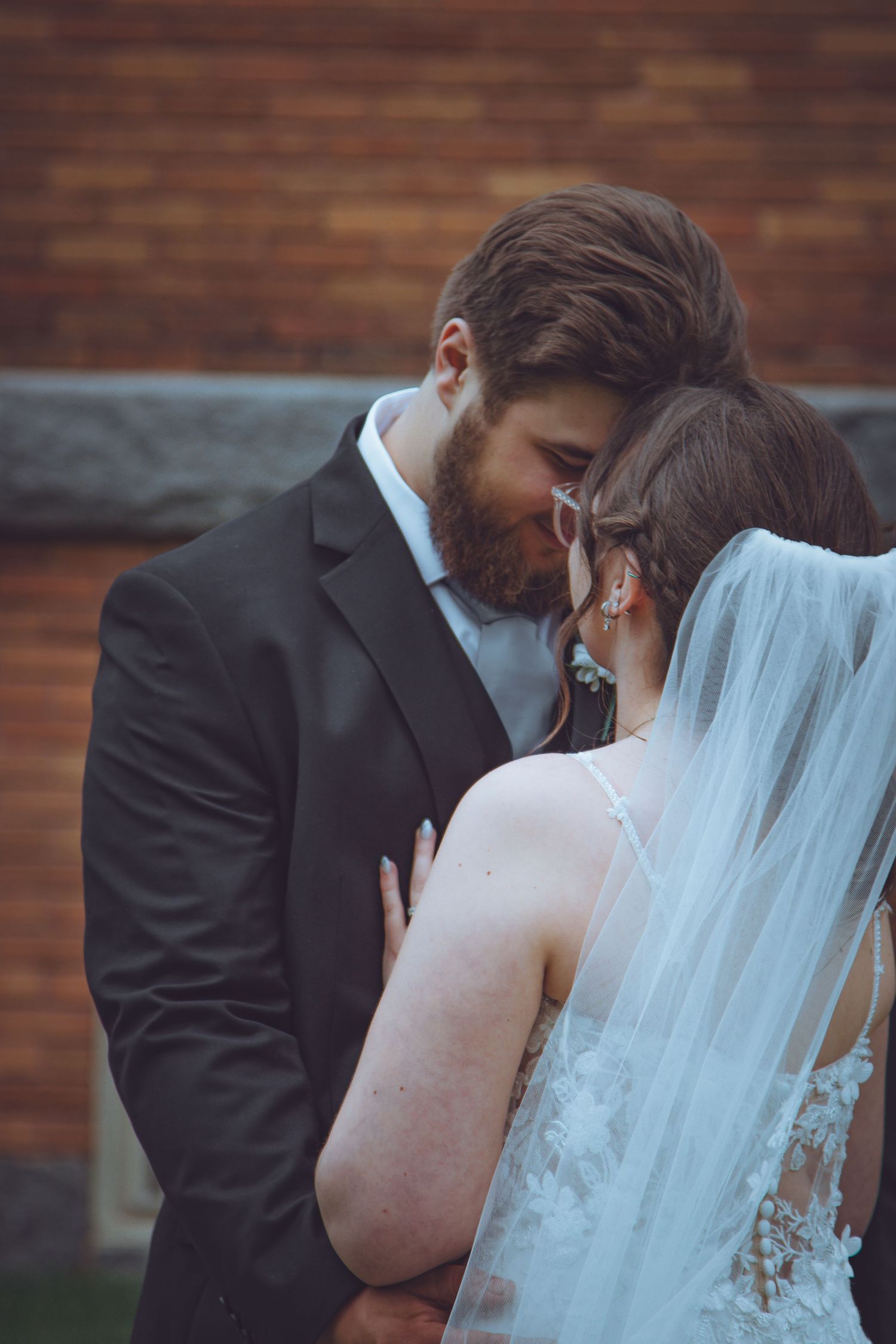 Intimate wedding moment between newlyweds as bride in flowing veil and groom in black suit share a tender embrace.