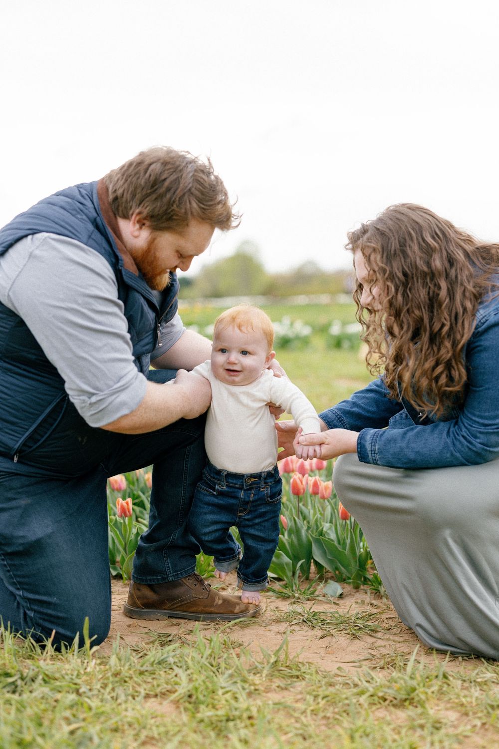 Two people in casual attire sit in a tulip field during springtime while interacting together in a sunny outdoor setting.
