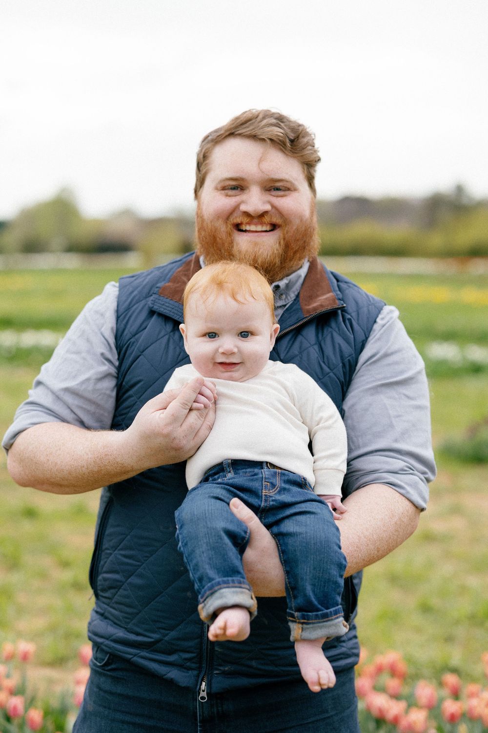 A parent and child sit together in a grassy field with pink flowers on a sunny spring day.