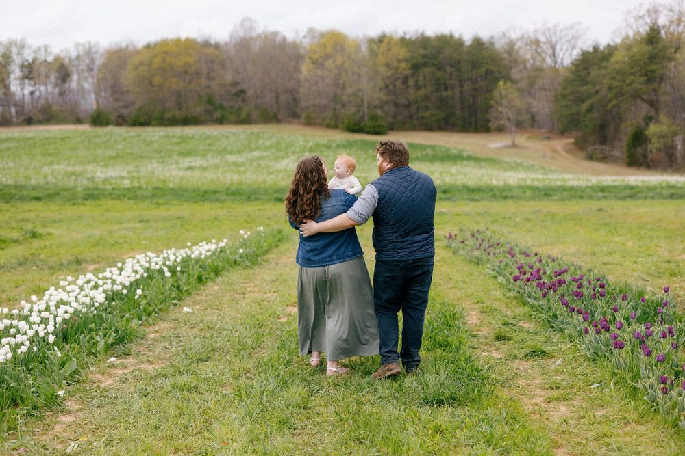 Three people walk through a green field with spring flowers near a dirt path on a sunny day.