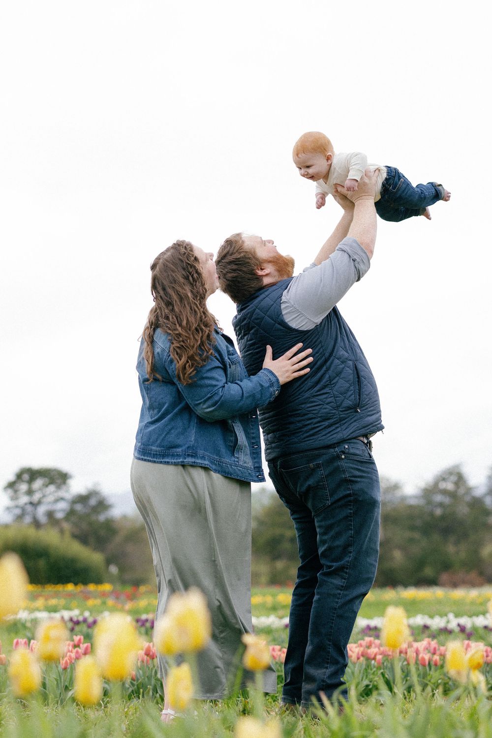 A couple shares joyful moments in a flower field while playing with a small child in matching blue jackets.