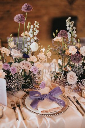 Elegant wedding table setting with purple allium flowers, pink roses, and white florals accented by a lavender napkin bow.