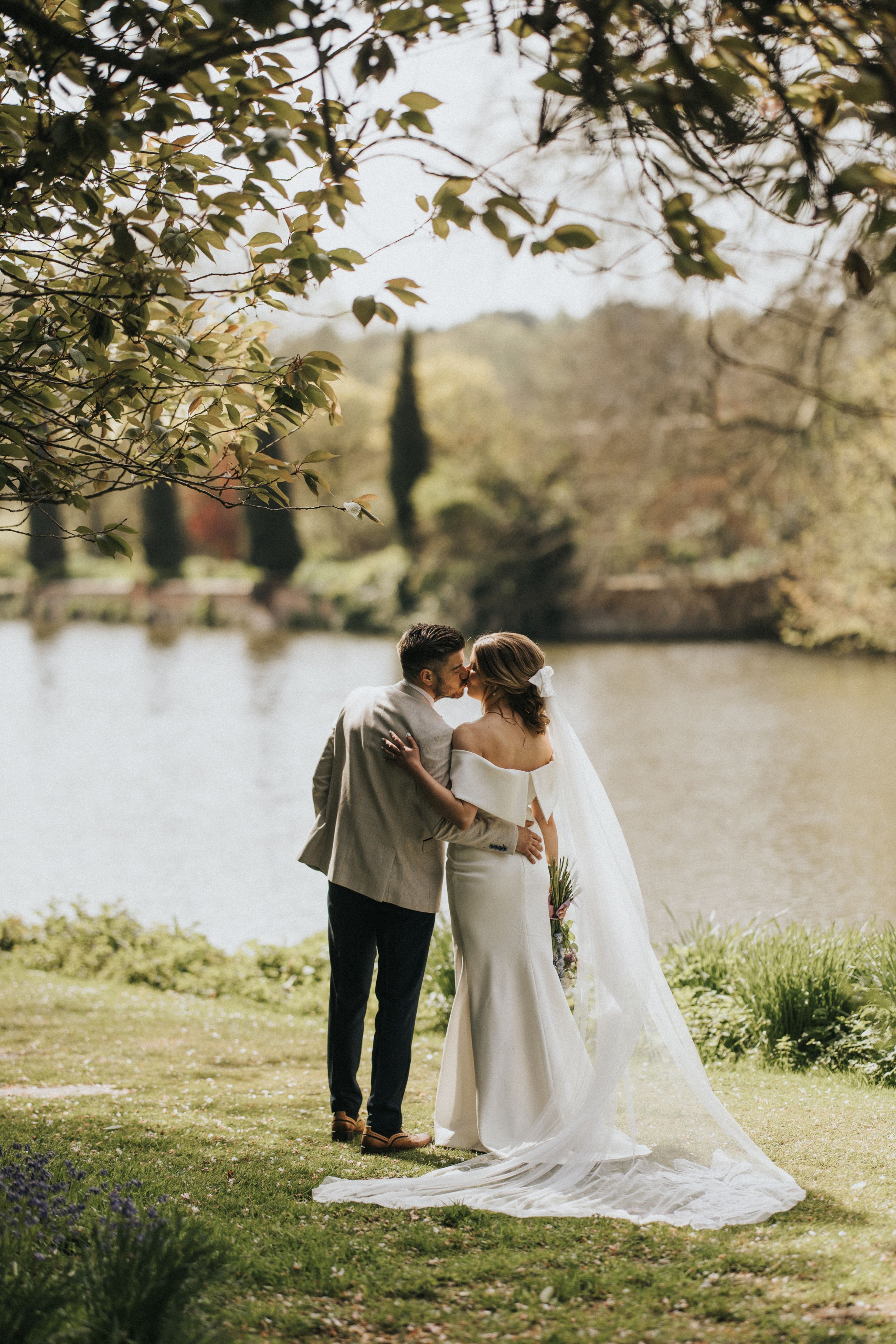 A romantic wedding couple shares a tender moment by a tranquil lake surrounded by lush greenery on their special day.