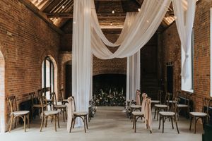 Indoor ceremony space with exposed brick walls, white draping fabric, and vintage wooden chairs arranged in rows.