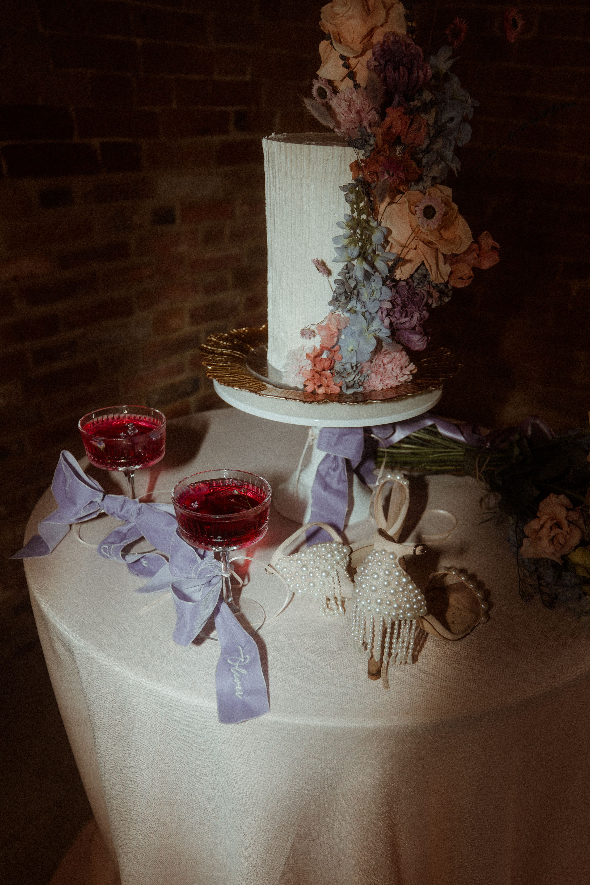 Rustic wedding cake decorated with cascading flowers next to red cocktails on white tablecloth against brick wall.