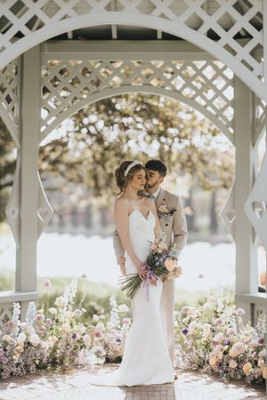Newlywed couple embraces under white garden gazebo surrounded by blooming pink and white flowers on sunny day.