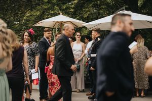 Guests mingle and socialize under white umbrellas at an outdoor event with lush greenery in the background.