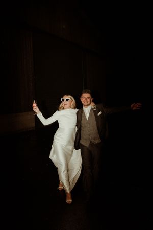 A couple dances together in a dark room, with the person on the left wearing sunglasses and a white dress.