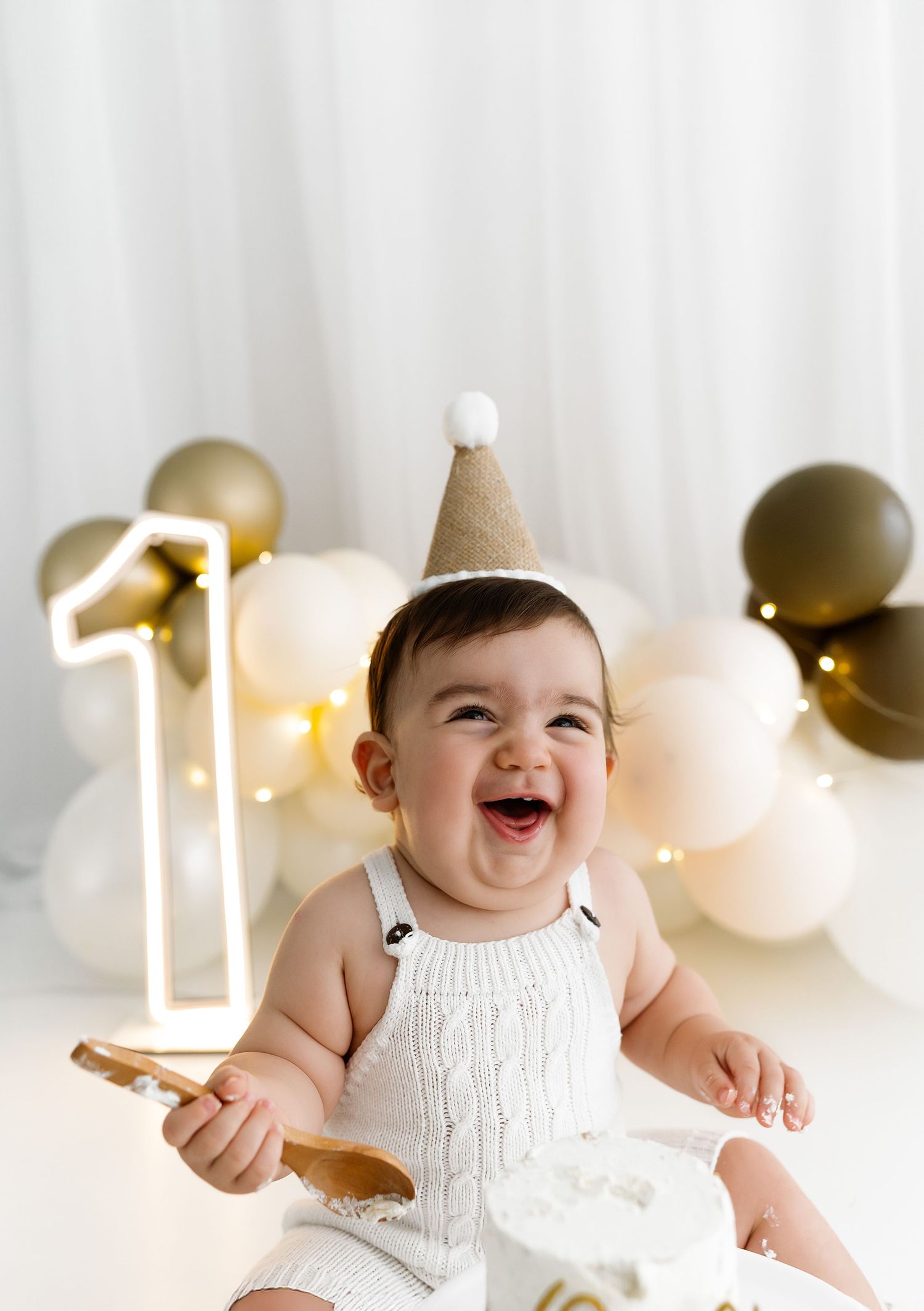 Baby wearing white romper and party hat celebrates first birthday with balloons and cake smash setup.