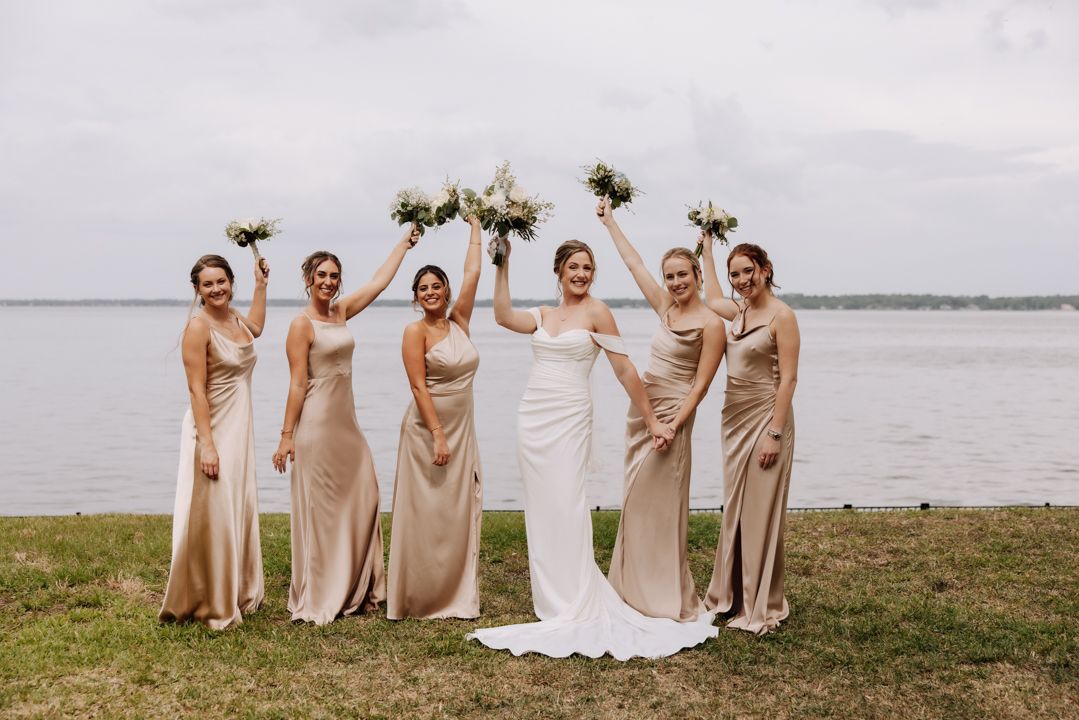 A bridal party in champagne colored gowns celebrates by a lakeside, with the bride in white raising her bouquet.