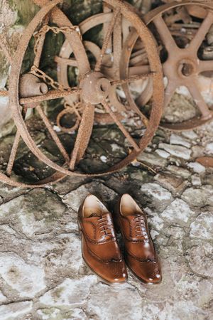 A pair of brown leather wingtip oxford dress shoes placed on stone with rustic metal wheels in the background.