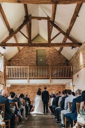 Rustic barn wedding ceremony with exposed wooden beams, brick walls and white vaulted ceiling in historic venue interior.