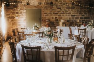 Rustic wedding reception venue with round tables, white linens and string lights in stone-walled room.
