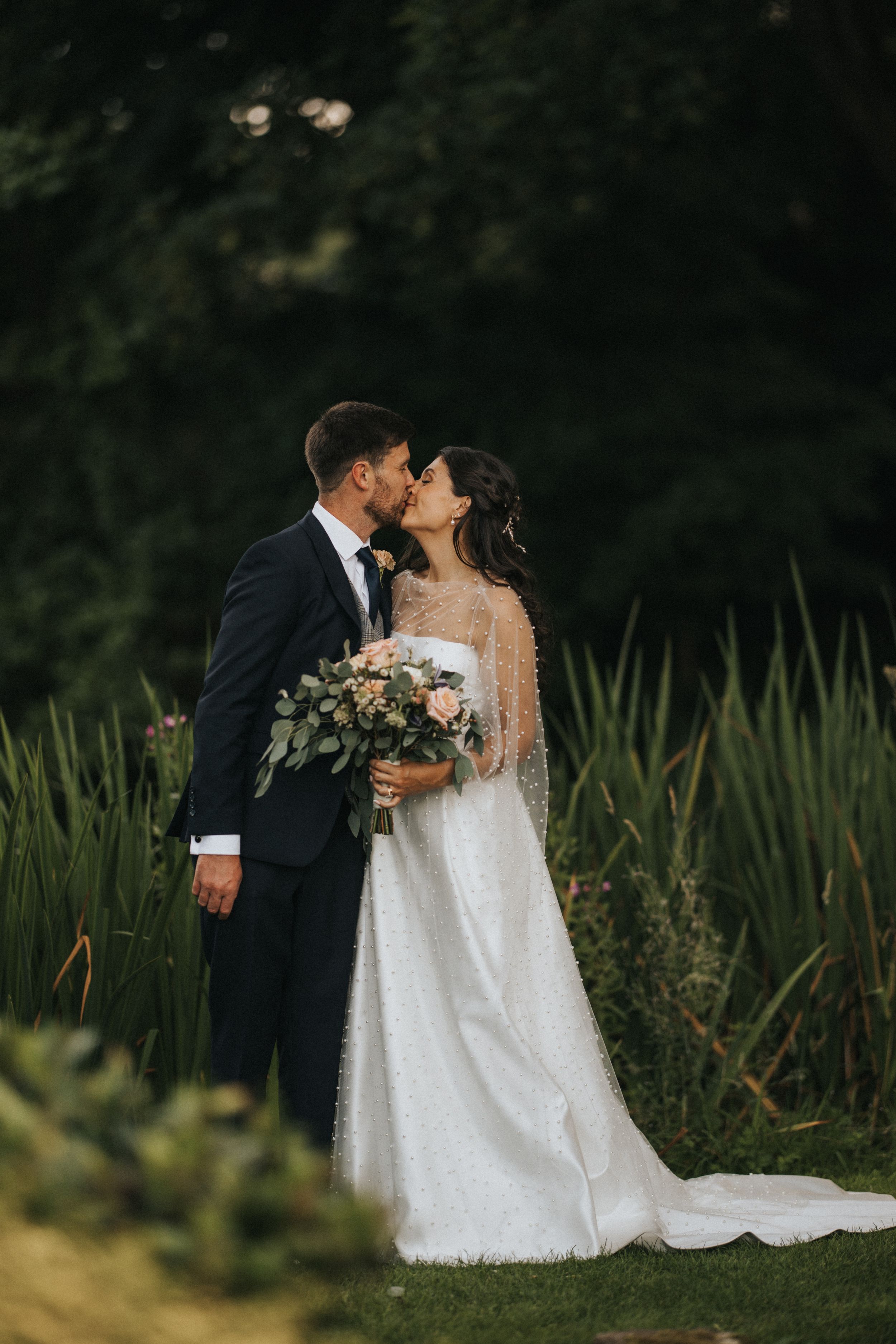A romantic newlywed couple shares a kiss in a garden setting with tall grass, while the bride holds a floral bouquet.