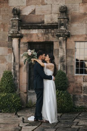 A couple embraces in front of a historic stone building entrance with ornate architectural details and greenery.