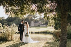 A couple in wedding attire embraces on a sunlit garden path near a tranquil pond surrounded by greenery.