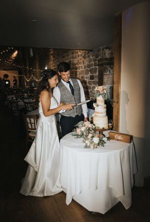 A couple stands together at their wedding reception by an elegant white cake decorated with fresh flowers and pink accents.
