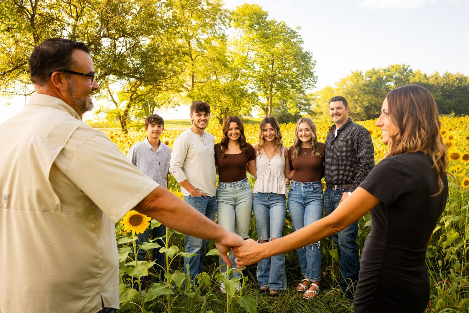 Group of people holding hands in prayer circle outdoors during golden hour with trees in background.