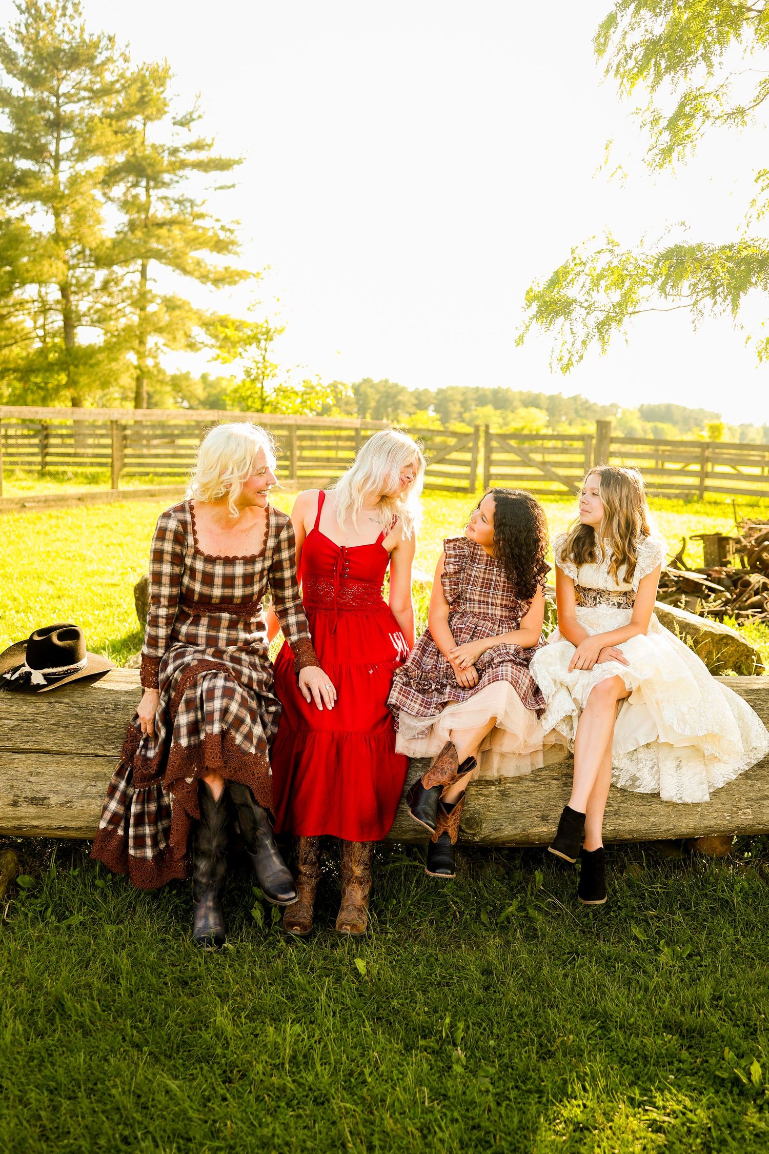 Four women in casual attire sit together on grass in a rustic countryside setting with sunlit trees in background.