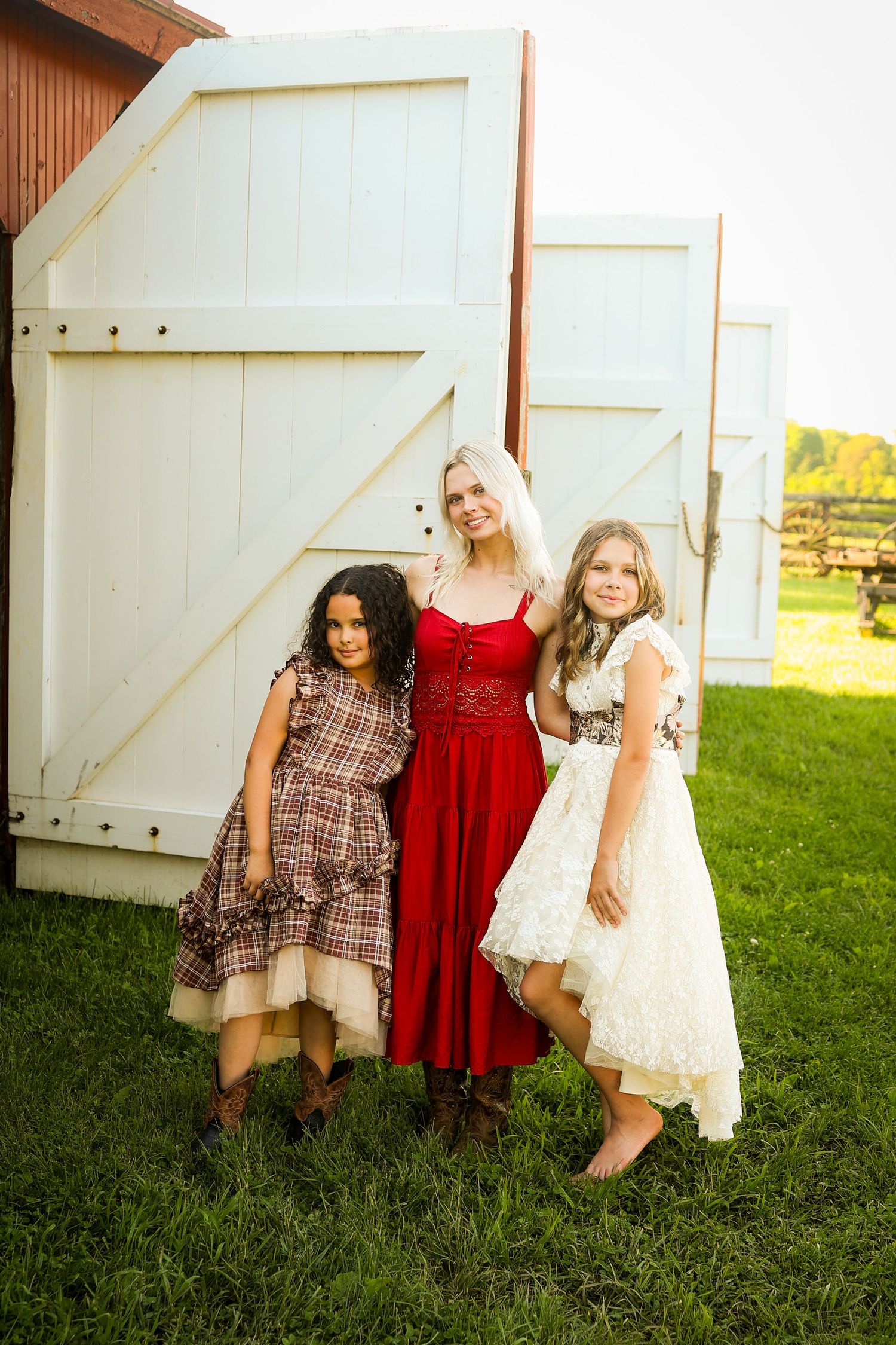 Three friends wearing dresses pose together in front of a white barn door in a grassy field.