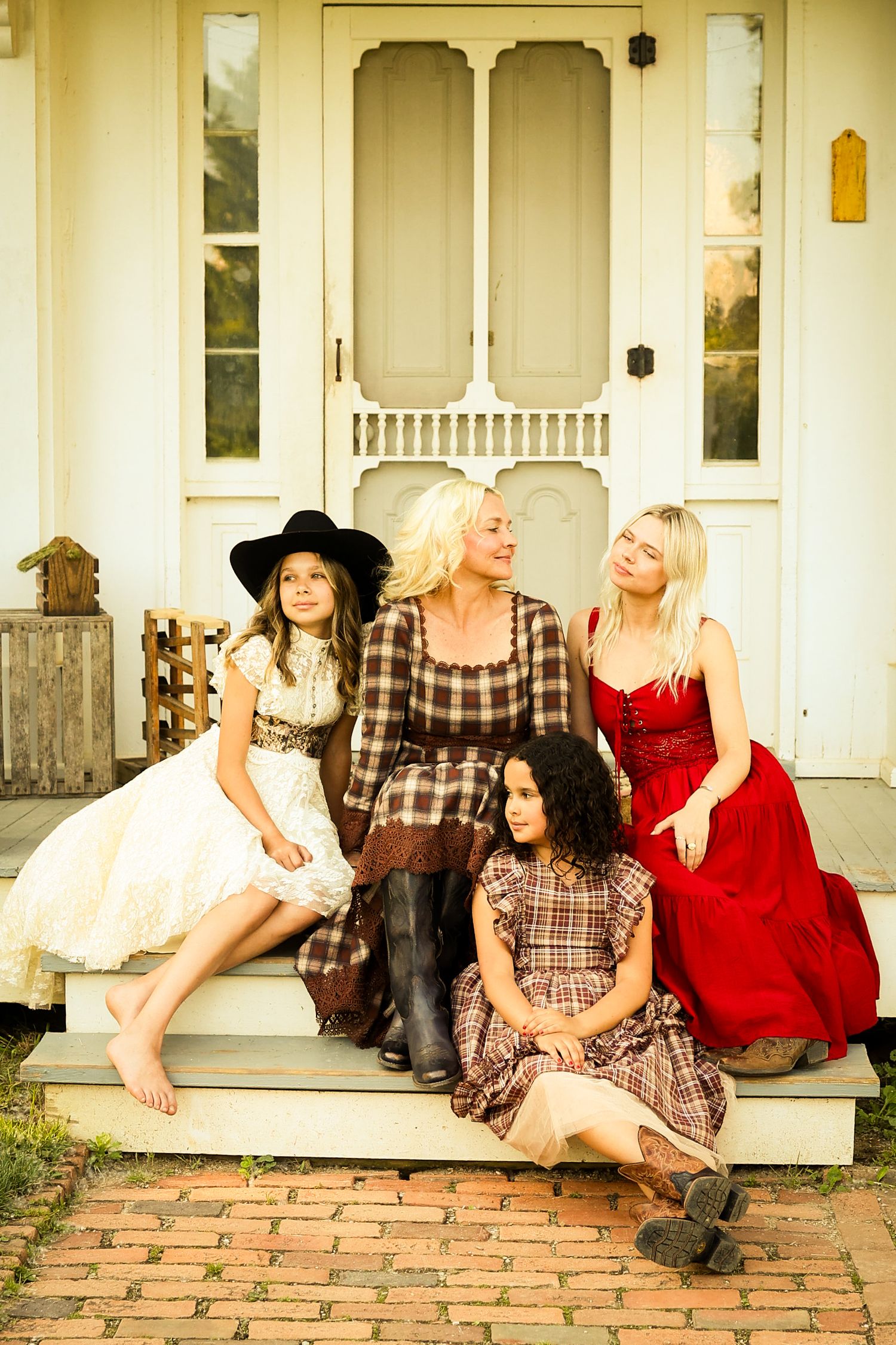 A stylish group poses on vintage porch steps wearing contrasting dresses in red, plaid, and white against a cream door.