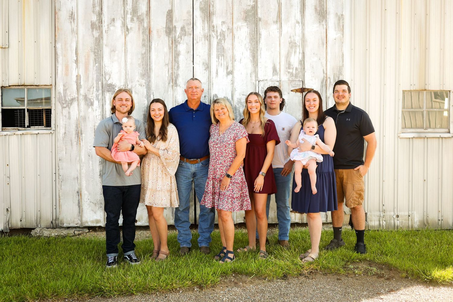 A family group poses together outside against a white barn wall on a sunny summer day.